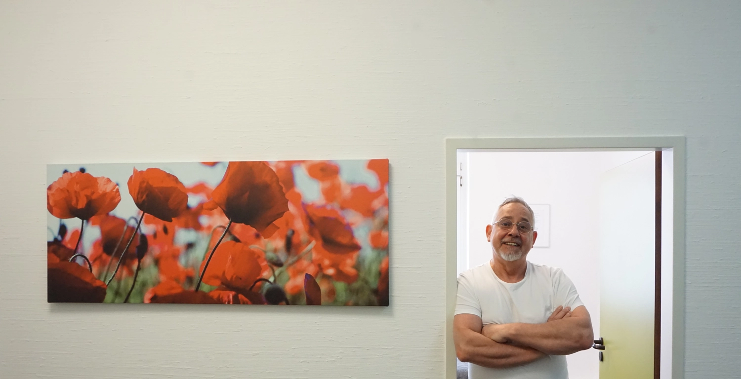 Praxis für Chiropraktik - Therapien und Behandlungs - Henry Burghammer steht mit verschränkten Armen in einem weißen T-Shirt in einem Türrahmen neben einem großen Panorama-Wandbild mit leuchtend roten Mohnblumen.